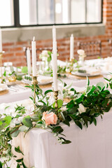 Long white tables set up for a wedding reception with greenery, roses, and tall candlesticks.