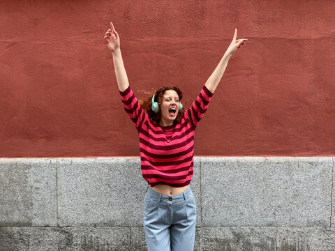 Joyful redhead woman dancing with headphones against red wall