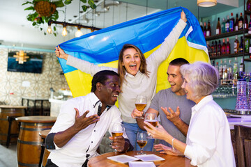 Cheerful diverse soccer supporters holding the flag of Ukraine and enjoing beer, spending time...