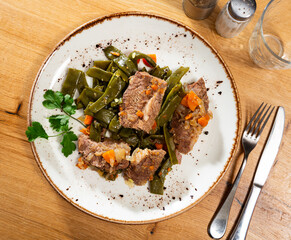 There is plate with braised beef fillet and stewed vegetable on wooden table. Stewed string beans...