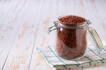 Raw red rice in glass storage jar on a white wooden table.