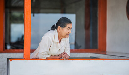 mujer mayor casual en su casa apoyada en un muro mirando para un lado con una leve sonrisa 