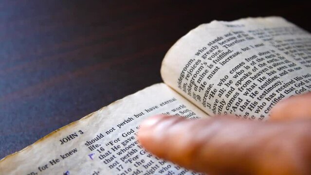 Hand pointing and gliding over John 3 verse 16 text on old bible page 1