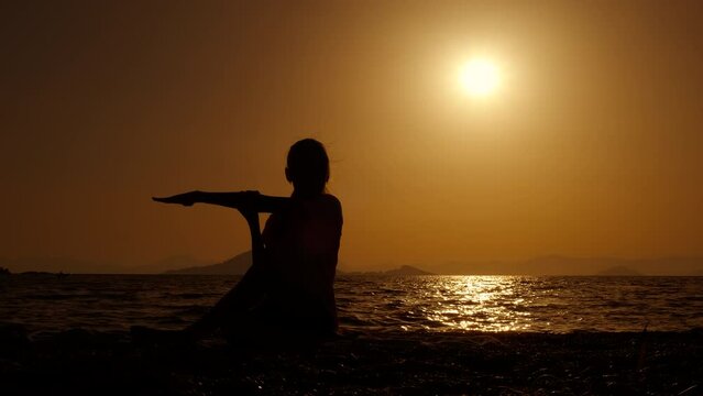 Teen silhouette relaxing with evening yoga. A girl silhouette relaxing and stretching her body in lotus pose against nightfall sky in summer.