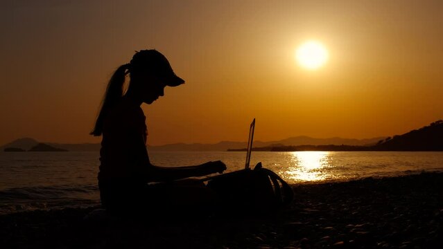 Teen luxury relaxation with notebook. A young girl silhouette relaxing with laptop on luxury evening seashore during weekend time.