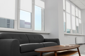 Comfortable sofa and table near large windows with white roller blinds indoors