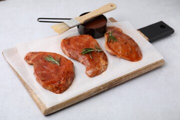 Board with raw marinated meat, rosemary and basting brush on light table, closeup