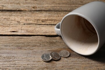 Poverty. Overturned dirty cup and coins on wooden table, closeup. Space for text