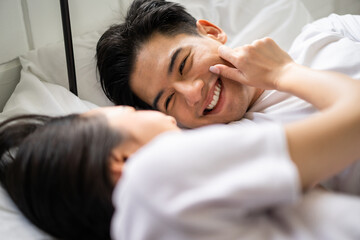 Asian new marriage couple lying down on bed and looking at each other.  - obrazy, fototapety, plakaty
