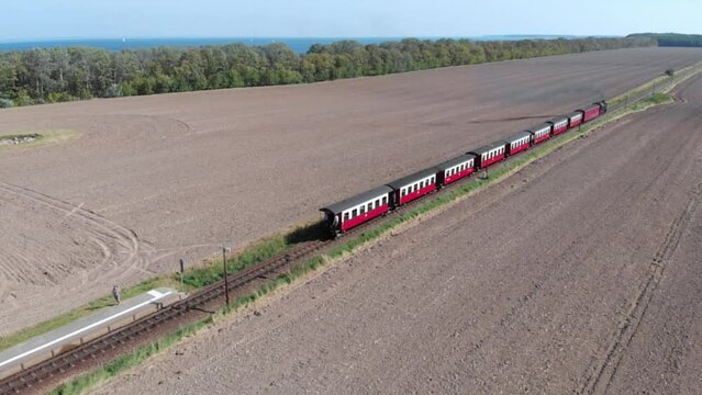 Aerial: steam narrow gauge railway in the countryside  passing by sown fields and with the sea in the background