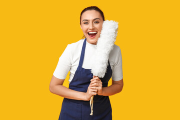Portrait of happy young housewife in apron with pp duster on yellow background