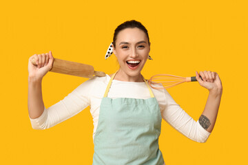 Portrait of happy young housewife in apron with whisk and rolling pin on yellow background