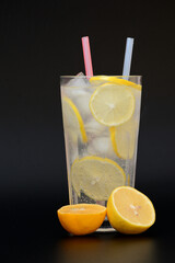 A tall faceted glass of refreshing lemonade with ice and mint on a black background, next to pieces of ripe citrus.