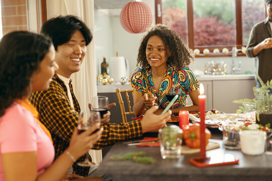Group of friends sitting around festive dinner table at home, eating and use mobile phone