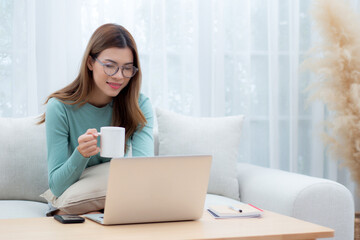 Young asian woman wearing glasses sitting sofa working on laptop computer and drinking coffee in living room at home, woman work from home with telework, freelance working, business and digital life.