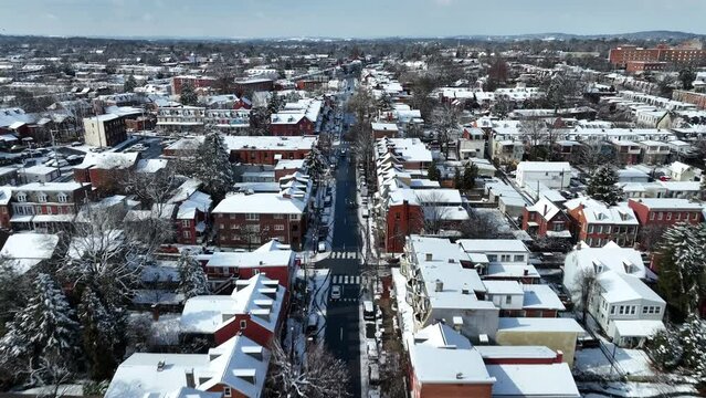 American city street with historic brick row houses covered in snow on sunny winter day. Aerial tracking shot above snowy roads, trees, and neighborhood.