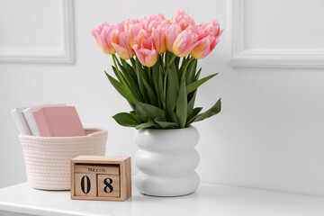 Calendar with date of International Women's Day, tulips and books on table near light wall