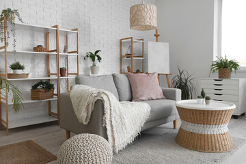 Loft style interior of light living room with cozy sofa, chest drawer, table, houseplants, shelving units and easel