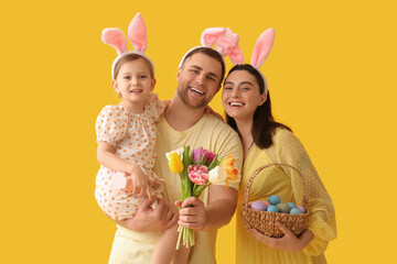 Happy family in bunny ears holding tulips and basket with Easter eggs on yellow background