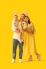 Happy family in bunny ears holding tulips and basket with Easter eggs on yellow background