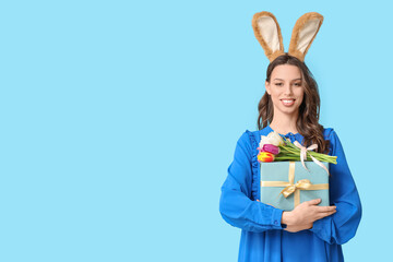 Happy young woman in Easter bunny ears headband with gift box and tulips on blue background