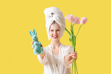 Happy young woman after shower holding Easter bunny and tulip flowers on yellow background