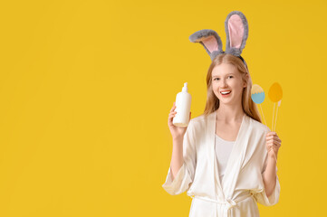Beautiful young woman in bunny ears holding cosmetic product and Easter decor on yellow background