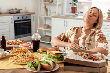 Beautiful woman at table full of unhealthy food in kitchen. Overeating concept