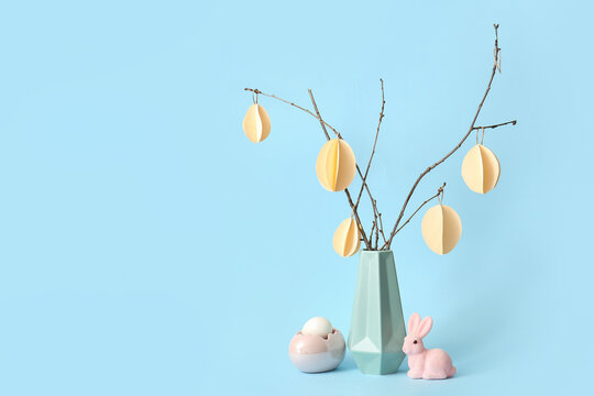 Branches with Easter paper eggs in vase, rabbit and egg holder on blue background