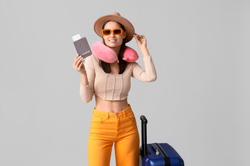 Young happy woman in hat and sunglasses with passport and suitcase on grey background