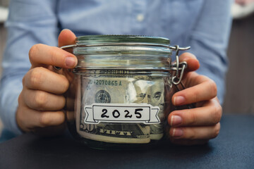 Unrecognizable woman holding Saving Money In Glass Jar filled with Dollars banknotes. 2025 year...