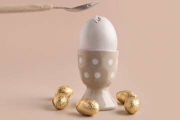 Teaspoon breaking egg in polka dot holder and chocolate candies on beige background, closeup....