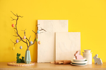 Vase with tree branches, Easter eggs, paintings and kitchen utensils on table near yellow wall