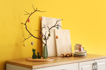 Vase with tree branches, Easter eggs, paintings and kitchen utensils on counters near yellow wall