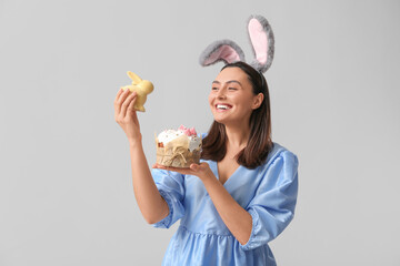 Beautiful young woman in bunny ears with sweet Easter cake and toy rabbit on grey background