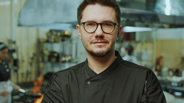 Young handsome professional chef in uniform and glasses standing in the restaurant kitchen and looking at camera with smile. Video portrait