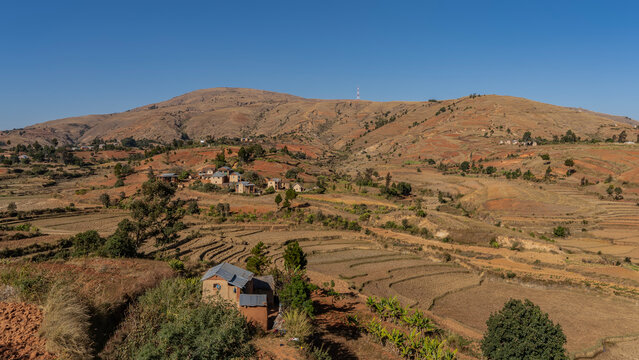 Rural landscape of Madagascar. Terraces of agricultural fields, houses on the slopes of the hill. Green vegetation on red earth. A mountain against a clear blue sky.