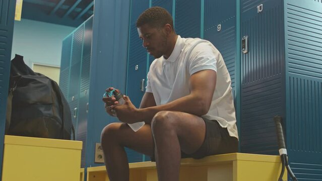 Slowmo shot of young Black male tennis player drinking water while sitting on bench in locker room before or after sports training