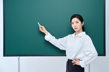 Wearing white heating, a beautiful Asian female teacher is holding a chalk in front of a green chalkboard and giving an Internet lecture with a confident expression and pose.