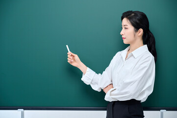 Wearing white heating, a beautiful Asian female teacher is holding a chalk in front of a green chalkboard and giving an Internet lecture with a confident expression and pose.