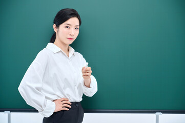 Wearing white heating, a beautiful Asian female teacher is holding a chalk in front of a green chalkboard and giving an Internet lecture with a confident expression and pose.