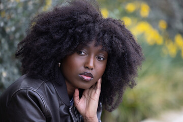 An African American woman with a stylish afro hairstyle gazes away thoughtfully. She is outdoors,...