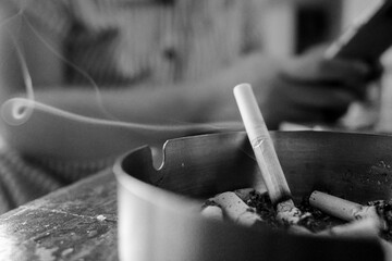 Burnt cigarette in an ashtray with a women on the background sitting while holding handphone in a...