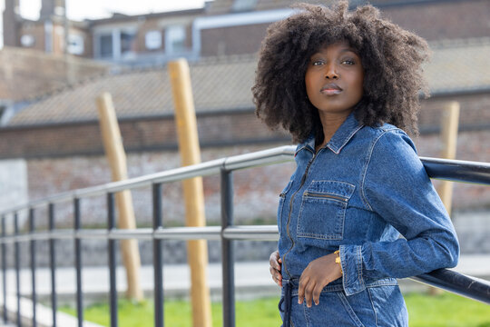 This image depicts a stylish African American woman with an impressive afro hairstyle standing confidently in an urban outdoor setting. She's wearing a denim outfit that radiates a casual yet chic