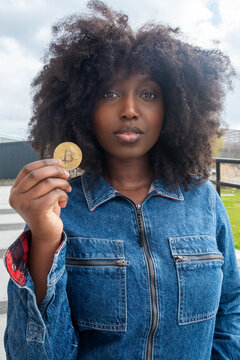 A young black afro american woman with natural, curly hair holds a Bitcoin token, her expression a mix of curiosity and skepticism. Dressed in a casual denim jacket, she stands outdoors, symbolizing