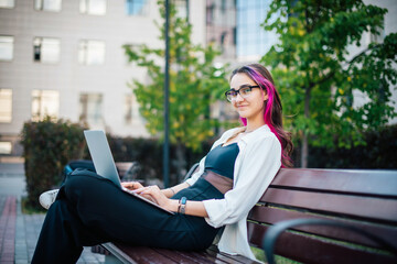 Portrait of a young beautiful female student sitting with a laptop on a bench outdoors. Long-haired...