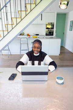 A bright and inviting image of a happy woman with braided hair, deeply engaged with her laptop at a spacious kitchen table. The modern kitchen background creates a comfortable setting that merges