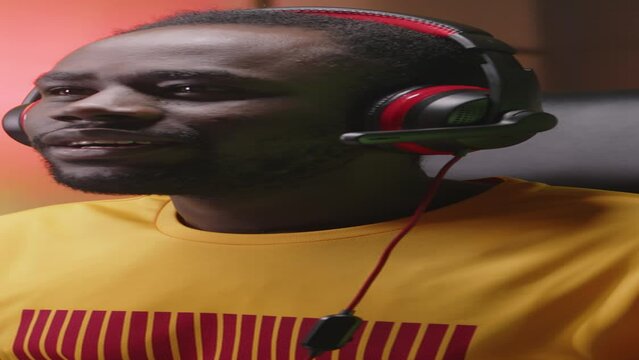 Vertical waist up of male African American gen z gamer wearing gaming headset with mic chatting with team players while playing personal computer game in his room with neon light and posters on walls
