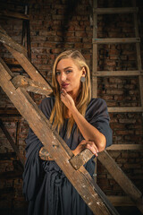 Beautiful girl in long gray dress posing in old rusty wooden step ladders on brick wall background. Beauty and fashion.