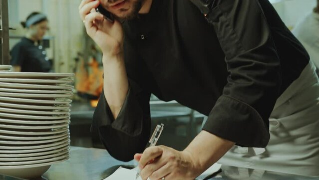 Male chef talking on mobile phone and writing down notes when taking order from customer during workday in restaurant kitchen